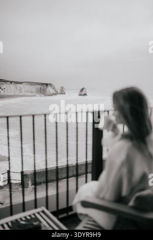 Woman sits on balcony, overlooking misty coastal cliffs. She sips coffee, calm and contemplative. Blurred motion suggests peaceful moment. Perfect for Stock Photo