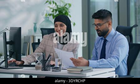 Two business colleagues happy positive in office talking with computer working Arabian businesswoman typing listening Muslim businessman leader boss Stock Photo