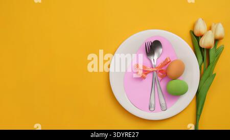 Easter Table Setting. Elegant plate with cutlery, eggs, and tulips on vibrant yellow background. Stock Photo