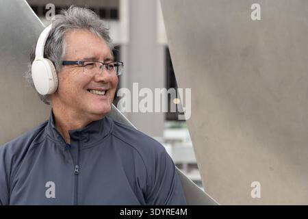 Senior man leaning on metal panels in urban plaza, wearing dark-gray jacket and white headphones Stock Photo
