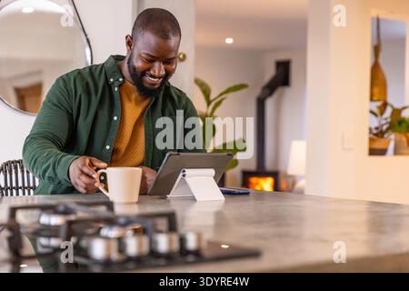 African American man smiling, using tablet on white adjustable stand at kitchen island, holding mug Stock Photo