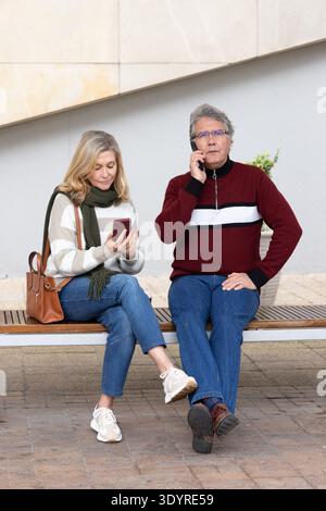 Senior couple sitting on bench at plaza, one checking red phone, other speaking on black phone Stock Photo