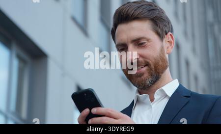 Close-up mature Caucasian business man holding mobile phone looking screen smiling in city businessman sending email with cellphone app using Stock Photo