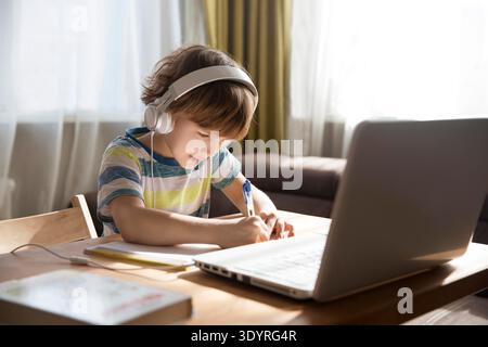 child boy in earphones is using a laptop and study online at home. homeschooling, distant learning || Model released Stock Photo