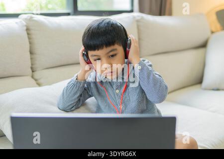 Happy Asian boy sitting on sofa using laptop and headphones in online class at home. Selective focus. Stock Photo