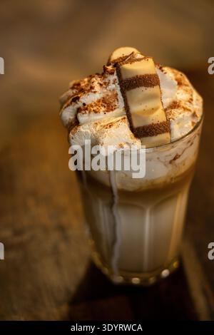 An editorial close-up of a gourmet iced coffee or latte served in a glass, topped with a large amount of whipped cream, cocoa powder, and a Kinder Bue Stock Photo
