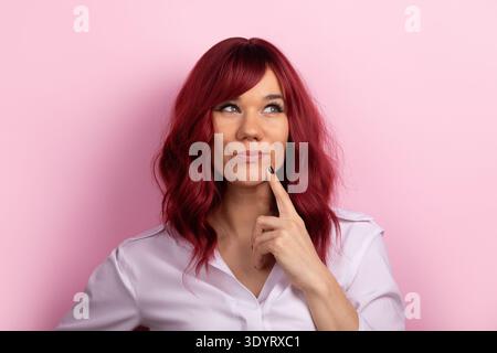 Portrait of a young woman with red hair in a white shirt, looking up and thinking, against a solid pink background. Stock Photo