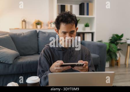 Man using smartphone while working on laptop at home workspace, representing remote work, digital communication and modern technology in everyday life Stock Photo