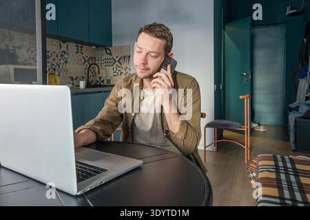 Man talking on smartphone while working on laptop at home office table. Male digital nomad on remote work, digital connectivity and managing tasks Stock Photo