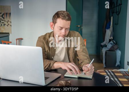 Man writing notes in notebook while working on laptop at home office table. Male digital nomad on remote work, planning and productivity in home Stock Photo