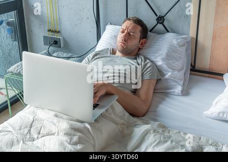 Man lying in bed working on laptop while resting with focused expression suggests concentration as he types and looks at the screen. Working from bed Stock Photo