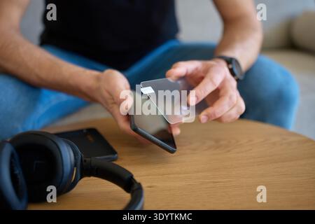 Man applies protective glass screen protector to smartphone on wooden table at home. Concept of device protection, mobile accessory installation and e Stock Photo