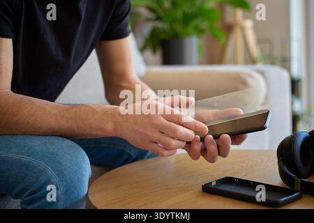 Man holding smartphone and applying protective glass screen protector while sitting by wooden table at home. Concept of device protection, mobile acce Stock Photo