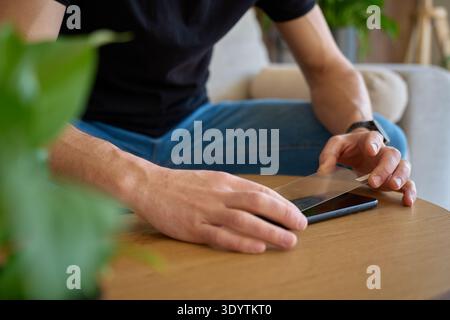 Man holding smartphone and applying protective glass screen protector while sitting by wooden table at home. Concept of device protection, mobile acce Stock Photo