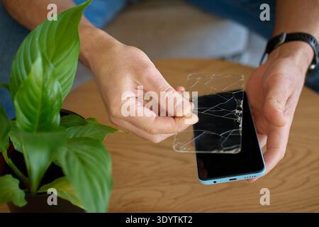 Man holds cracked protective glass screen protector above smartphone on wooden table. Concept of device repair, screen protector replacement and mobil Stock Photo