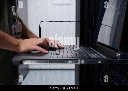 Data center technician typing on a laptop console while monitoring rack servers during system maintenance Stock Photo