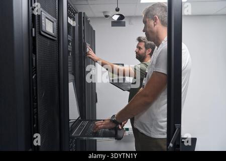 Two IT technicians troubleshooting servers in a secure data center server room with laptops Stock Photo
