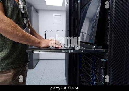 It technician working on a laptop console in a data center server rack performing network maintenance and monitoring Stock Photo
