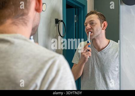 Man brushing teeth with electric toothbrush while looking into bathroom mirror. Daily hygiene routine and morning self care. Oral health and dental Stock Photo