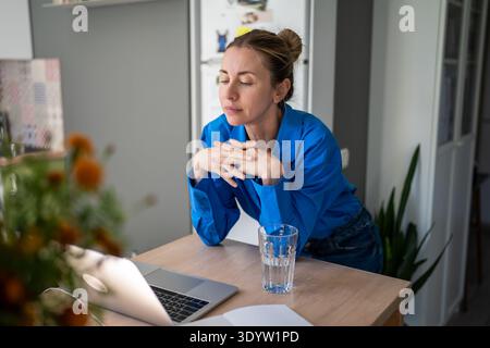 Thoughtful positive woman watching popular culinary program online on laptop, learning new recipes. Stock Photo