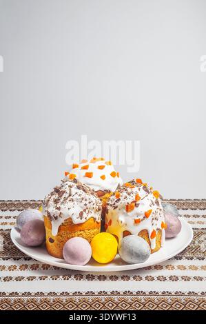 A collection of beautifully decorated Easter cakes and colorful eggs are presented on a white plate, set against a backdrop of a traditional embroider Stock Photo