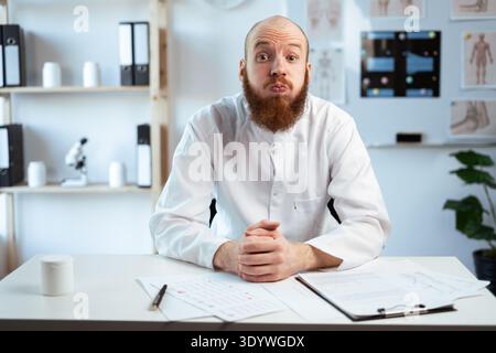 Bearded male doctor with prescriptions, medication documents for patients holds breath blows cheeks makes funny grimace works in hospital office. copy Stock Photo