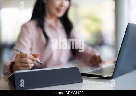 Asian adult woman using modern laptop and digital pen on a tablet at her workspace. She is focused on creative design or business tasks. Stock Photo