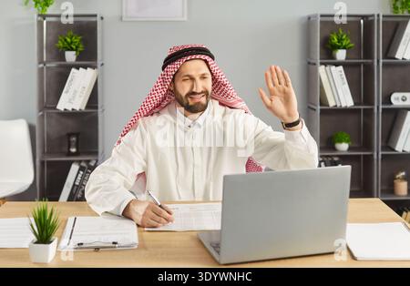 Young Arab businessman sitting at the desk waving hand having video call on a laptop in office. Stock Photo