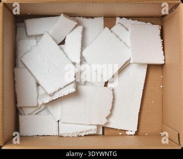 broken white styrofoam pieces in brown corrugated cardboard box, flat polystyrene foam boards with irregular shapes in top view, packaging waste Stock Photo
