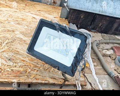 Water flows over a damaged LED light fixture on a construction site. The fixture sits on wooden boards, showing signs of neglect. Stock Photo