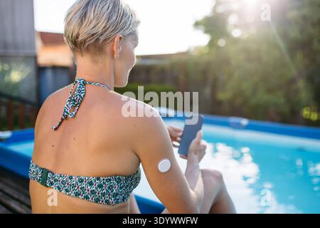 Woman using smartphone with wearable health sensor on arm. Woman using smartphone with wearable health sensor on arm by poolside. model released Stock Photo