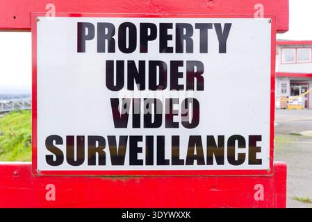 A security sign stating "PROPERTY UNDER VIDEO SURVEILLANCE" mounted on a bright red wooden fence near a harbour building. Stock Photo