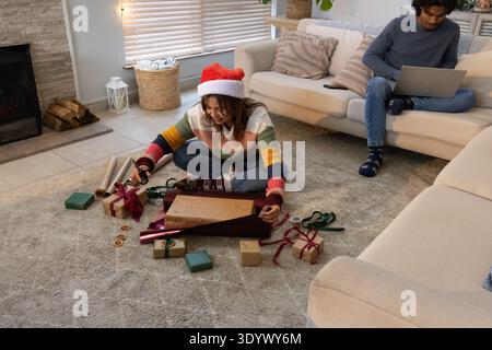 Couple wrapping in living-room woman in Santa-hat cutting paper on rug man using laptop, copy space Stock Photo