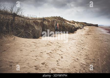 Deserted beach with sand dunes pushing and slipping against a chain link fence under a cloudy sky. Stock Photo