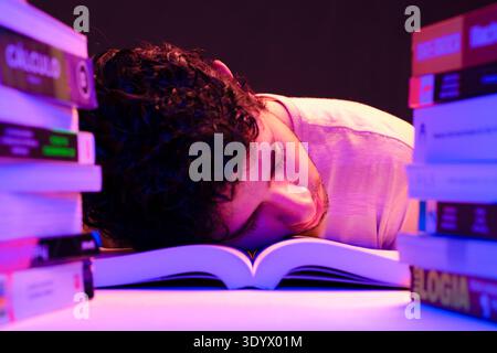 A young man, tired from studying, falls asleep on top of books in his room with the light on, sitting and studying. Stock Photo