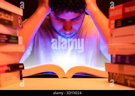 Guy with hand on his head, studying among books in his room with light coming from the book, sitting focused on his reading. Stock Photo
