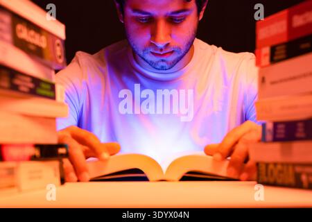 Young man studying among books in his room with light emanating from the book, sitting focused on his reading Stock Photo