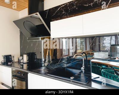 Modern kitchen with black marble countertop, white cabinets, stylish hood, coffee machine and black sink in minimalist luxury interior. High quality p Stock Photo