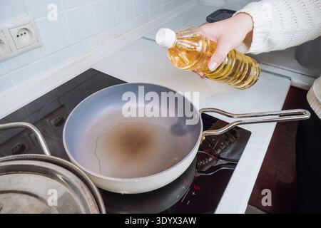 Woman pouring yellow vegetable oil from a plastic bottle into a frying pan on a modern induction stove to prepare a meal in a home kitchen Stock Photo