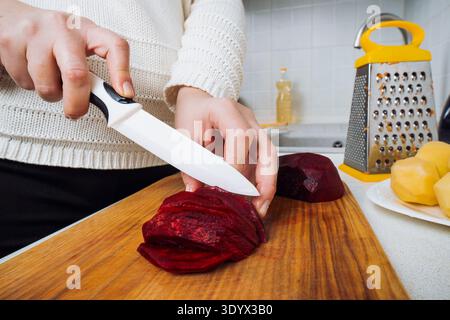 Close up of female hands slicing peeled red beetroot with a white knife on a wooden board to cook traditional Ukrainian borscht in a home kitchen Stock Photo