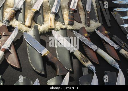 Selection of handcrafted hunting knives with engraved steel blades and decorative handles displayed with leather sheaths at an outdoor artisan craft m Stock Photo