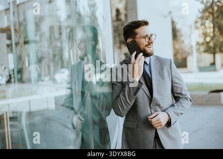 A man in a suit is standing outside a glass building. He is smiling and talking on his phone. The sun is shining and the area is busy. A reflection is Stock Photo