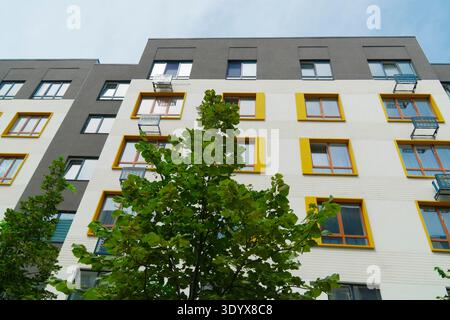 Low angle view of a modern residential building facade featuring bright yellow window frames, white and grey walls, and green trees in front Stock Photo