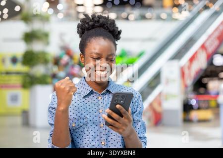 Happy young woman in a busy shopping mall celebrates good news on her smartphone, smiling and pumping a fist in triumph while browsing stores and using a mobile app Stock Photo