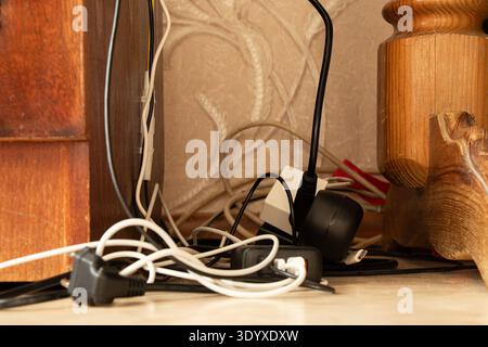 Tangled cables, plugs, and chargers lie on the floor near an outlet and surge protector behind furniture and are used for the following themes: wire c Stock Photo