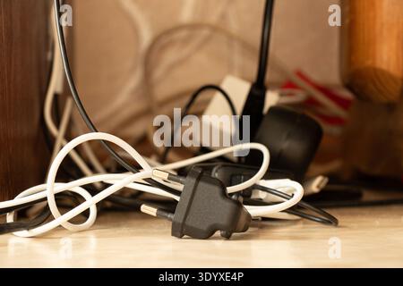 Tangled cables, plugs, and chargers lie on the floor near an outlet and surge protector behind furniture and are used for the following themes: wire c Stock Photo