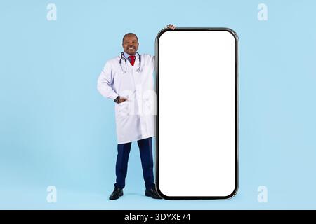 Black doctor standing next to oversized smartphone mockup on blue studio background Stock Photo