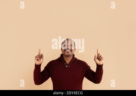 Black man pointing upward with hand gesture while smiling on beige background Stock Photo