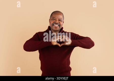 Smiling mature black man wearing burgundy sweater making heart shape with hands Stock Photo