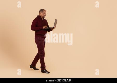 Mature black man walking while using laptop on beige studio background Stock Photo